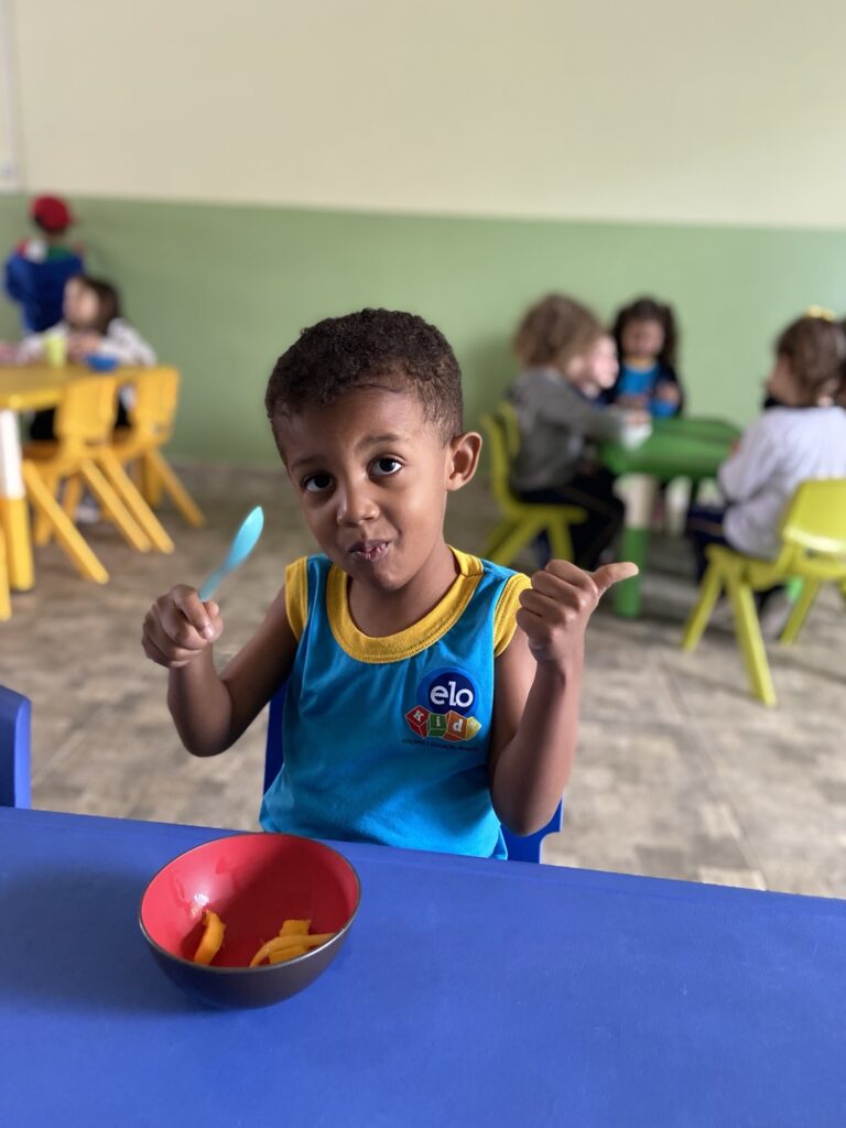 Criança sorrindo com colher na mão durante refeição, uniforme da Elo Kids, turma ao fundo