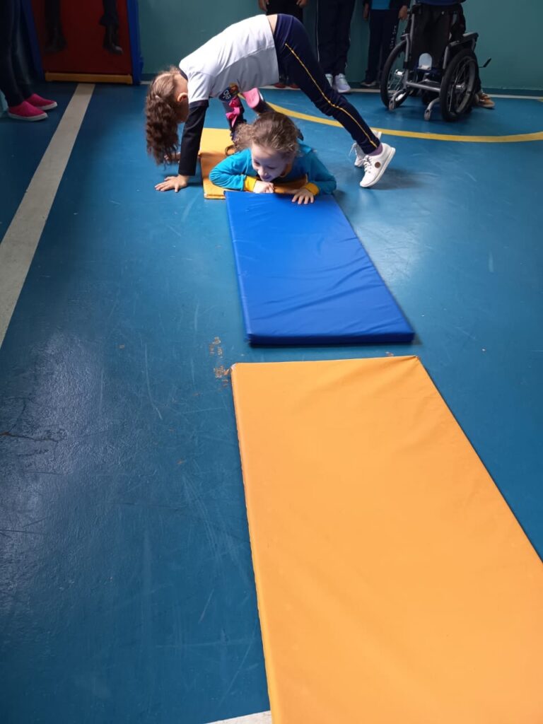 Professora auxiliando criança em exercício de alongamento na aula de educação física